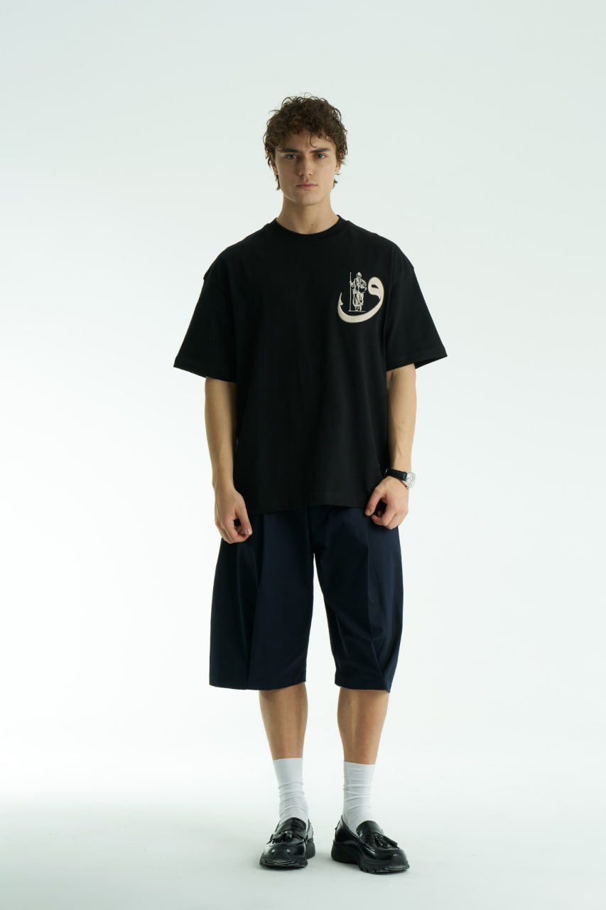 Black Oversized cotton graphic T-shirt with small chest emblem and large ornate framed back print, shown in black, white, brown, and navy.