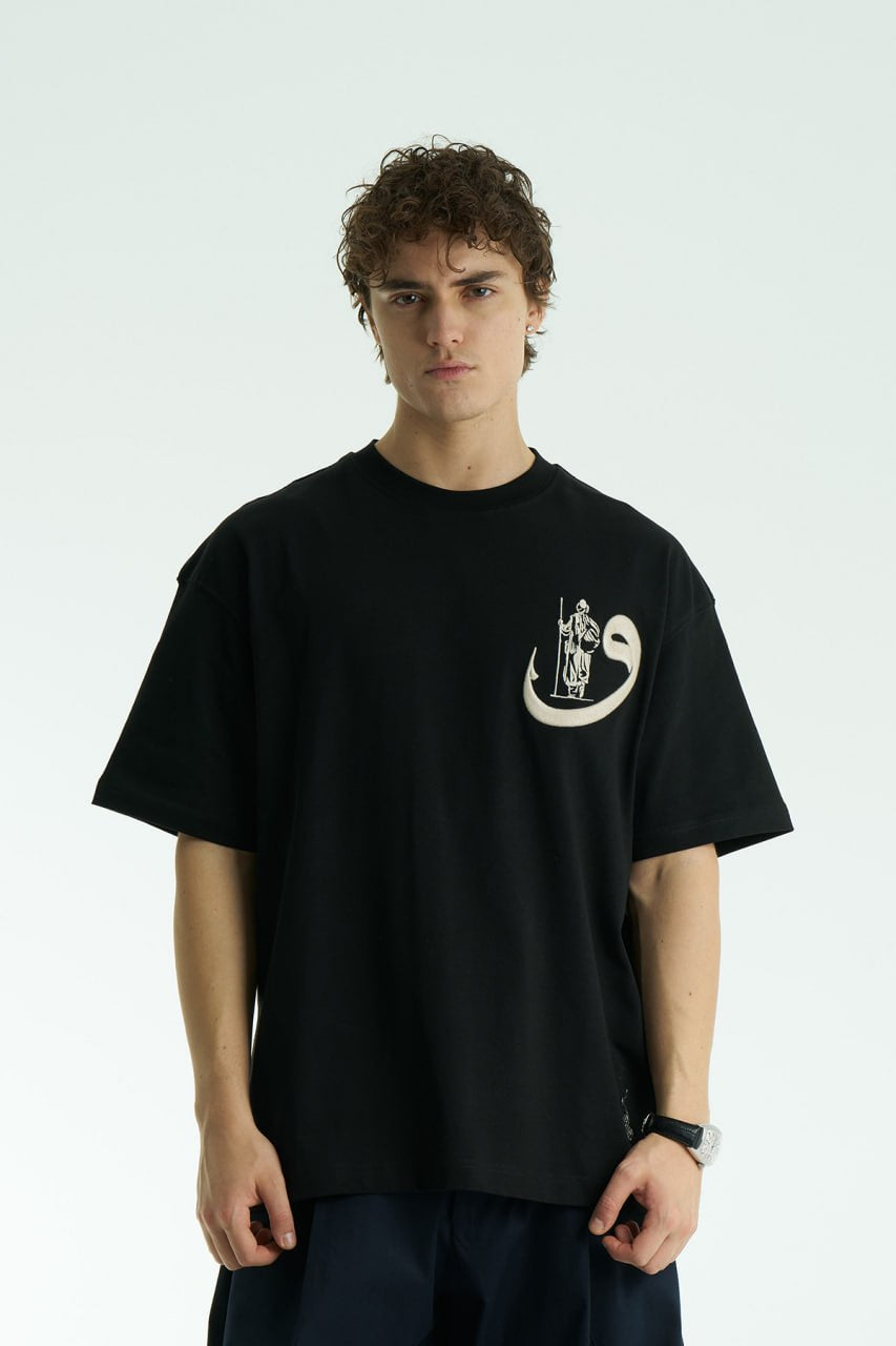 Black Oversized cotton graphic T-shirt with small chest emblem and large ornate framed back print, shown in black, white, brown, and navy.
